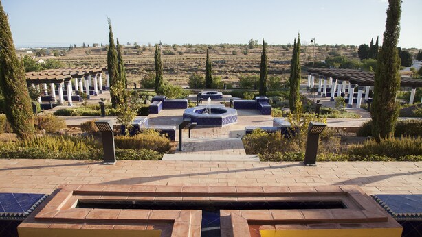 Une cour intérieure avec une fontaine centrale, entourée d’espaces aménagés pour s’asseoir et de grands cyprès.