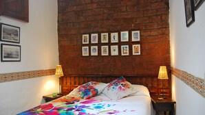 Une chambre avec un mur en briques, un lit recouvert d’une literie aux motifs floraux colorés, deux lampes de chevet et une série de tableaux encadrés accrochés au mur.