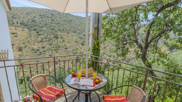 A balcony with wicker chairs, a table with a book and fruit, and a view of a hilly landscape.