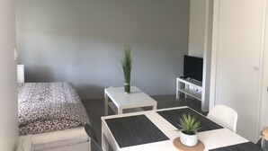 A modern, minimalist room with a bed, a small table with a plant, a TV on a stand, and a dining area with a table and chair. - House 4 people near sea (Les Sables-d'Olonne)