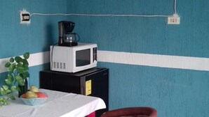 Ein Zimmer mit blauer Wand, einem weißen Tisch mit weißem Tuch, zwei Korbstühlen, einem kleinen schwarzen Gerät auf dem Tisch und einer Steckdose an der Wand.