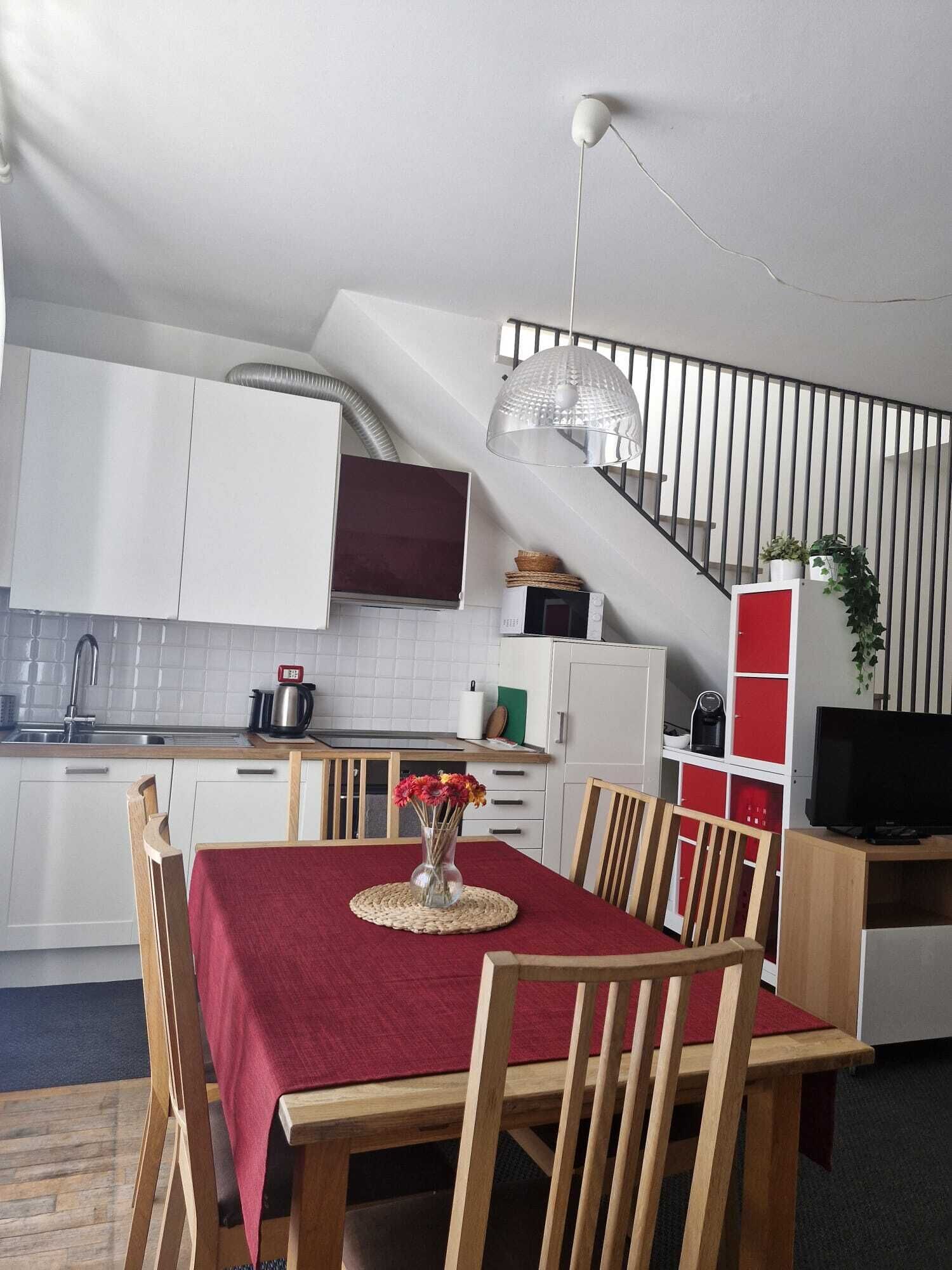 A modern kitchen with white cabinets, a dining table with a red tablecloth, and a staircase with a glass railing.