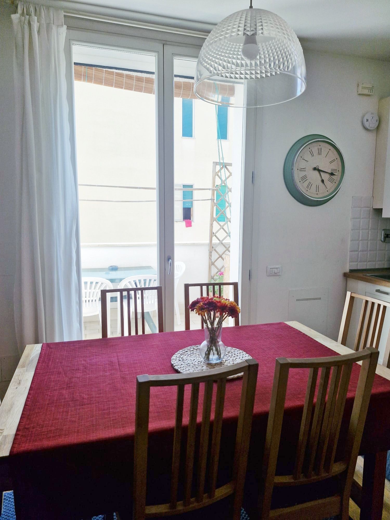 Uma área de refeições com uma mesa posta para quatro pessoas, um relógio na parede e vista através de uma porta de vidro.
