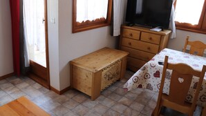 Una habitación pequeña con un banco de madera, un televisor sobre un aparador, una mesa con mantel estampado y una silla.