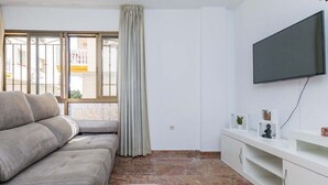 Un salon comprenant un canapé, un téléviseur fixé au mur, une fenêtre avec des rideaux et un meuble blanc agrémenté d’objets décoratifs.