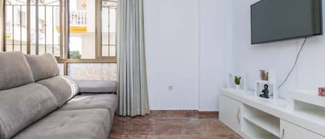 A living room with a sofa, a TV mounted on the wall, a window with curtains, and a white cabinet with decorative items.