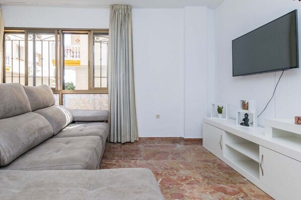 A living room with a sofa, a TV mounted on the wall, a window with curtains, and a white cabinet with decorative items.