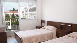 A bedroom with two beds, a wooden nightstand, a chair, and a window with a view of buildings and trees.