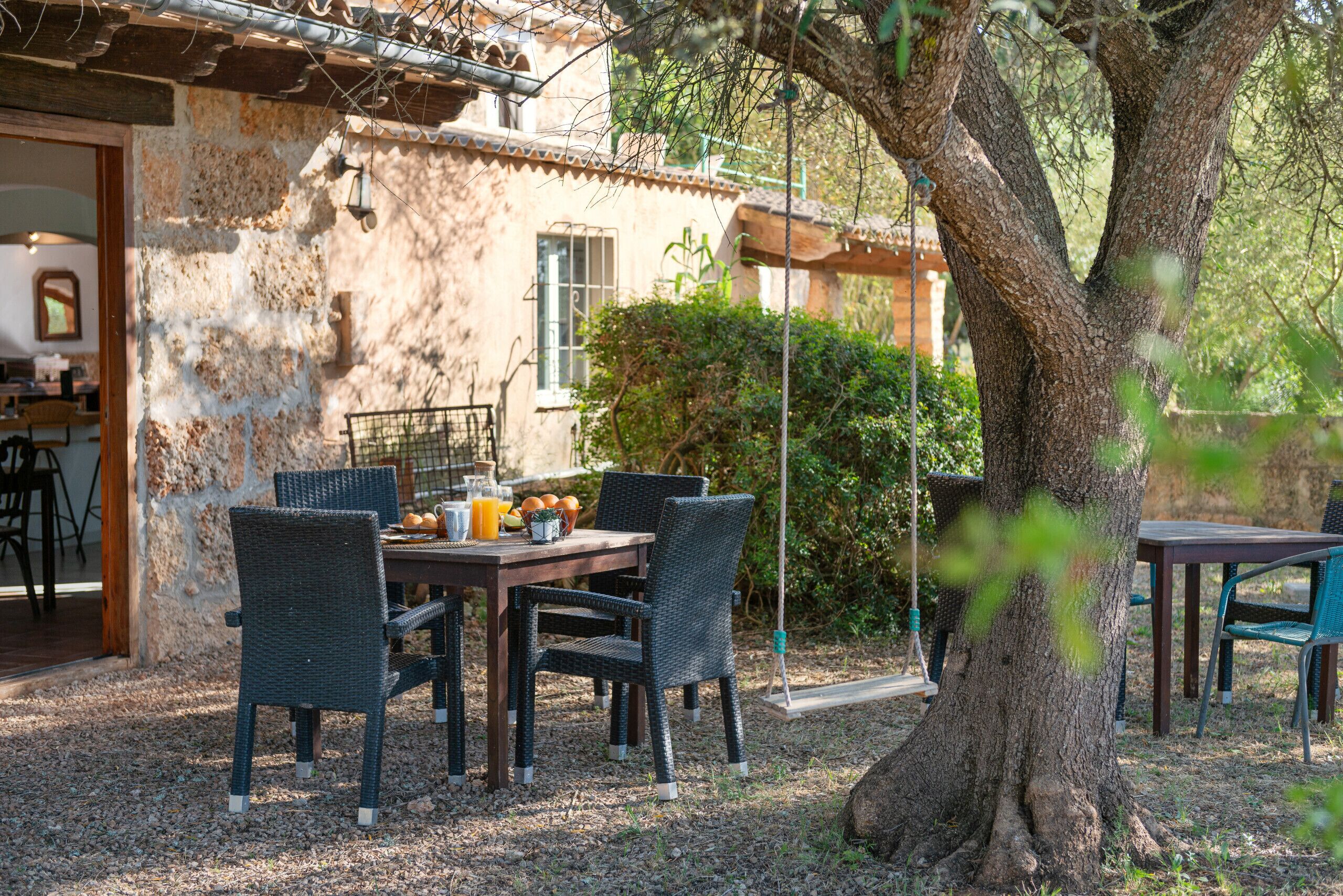 An outdoor dining area with wicker chairs and a table set with fruits and drinks, surrounded by a rustic stone building and a large tree.