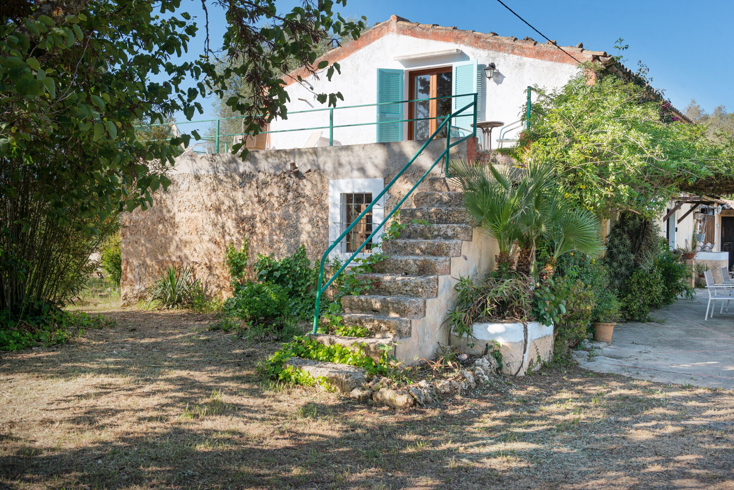 Une maison rustique avec un escalier en pierre menant à une porte en bois, entourée de verdure et d’un mur de pierres.
