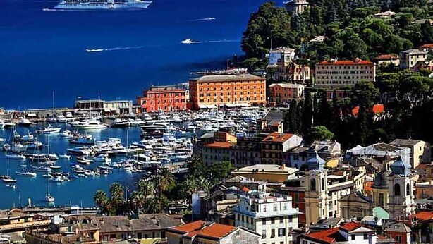 Un pueblo costero con un puerto deportivo lleno de barcos, edificios históricos y un crucero al fondo.