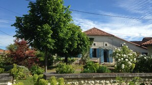 Ein traditionelles Haus mit einer Steinwand im Vordergrund, ein großer Baum und ein Garten mit verschiedenen Pflanzen und Blumen.