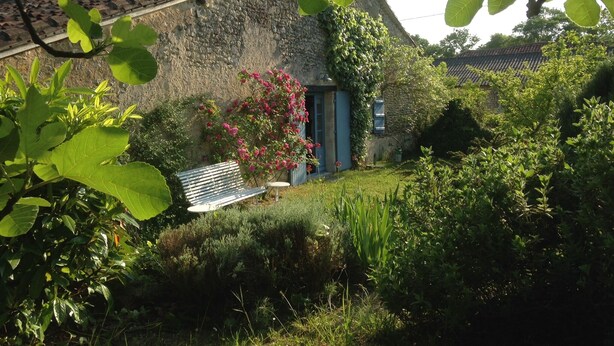 Ein rustikales Steinhaus mit Strohdach, einer Holzbank und einem Garten mit verschiedenen Pflanzen und Blumen.
