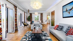 A modern living room with a sofa, coffee table, and chairs. There is a large window with curtains, a hanging light fixture, and a painting on the wall. - ❀Family Paradise. Peaceful, golf, countryside & beach, pool, close to everything (Lagos)