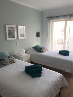 Two single beds with white bedspreads and teal pillows, set against a light blue wall with framed artwork.