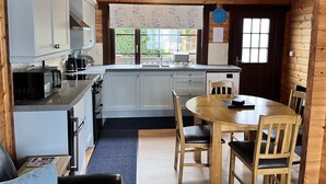 A compact kitchen with white cabinets, a sink, and a dining area with a wooden table and chairs. - Willow Lodge, Holiday Log Cabin, Cedar Springs, Heacham. Close to the beach. (Heacham)
