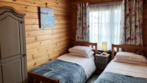 A wooden cabin room with two beds, a nightstand, a lamp, and a painting on the wall. - Willow Lodge, Holiday Log Cabin, Cedar Springs, Heacham. Close to the beach. (Heacham)