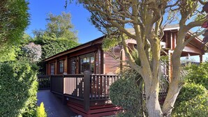 A wooden cabin with a deck, surrounded by lush greenery and trees. - Willow Lodge, Holiday Log Cabin, Cedar Springs, Heacham. Close to the beach. (Heacham)
