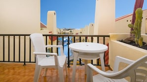 Un balcon avec des chaises en plastique blanches et une petite table ronde, donnant sur une piscine et un bâtiment avec un toit rouge.