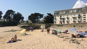 A beach with people sunbathing and sitting under umbrellas, a building with balconies, and trees in the background.