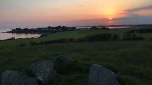A sunset over a grassy field with rocks in the foreground and a distant shoreline with buildings.