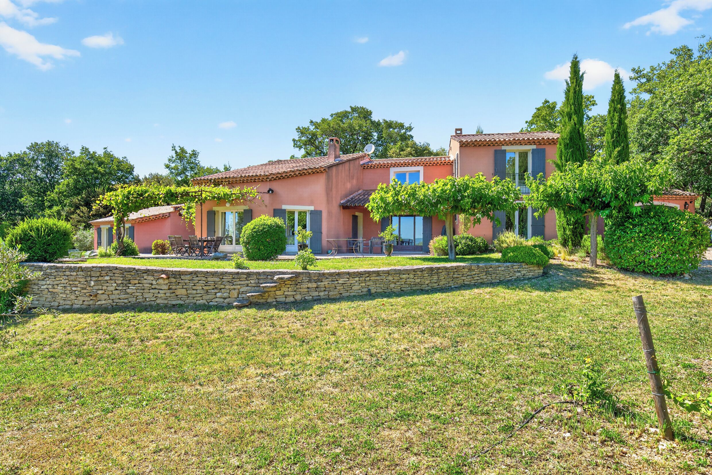 Une maison à deux étages avec un mur en pierre, un jardin et un ciel d’un bleu éclatant.