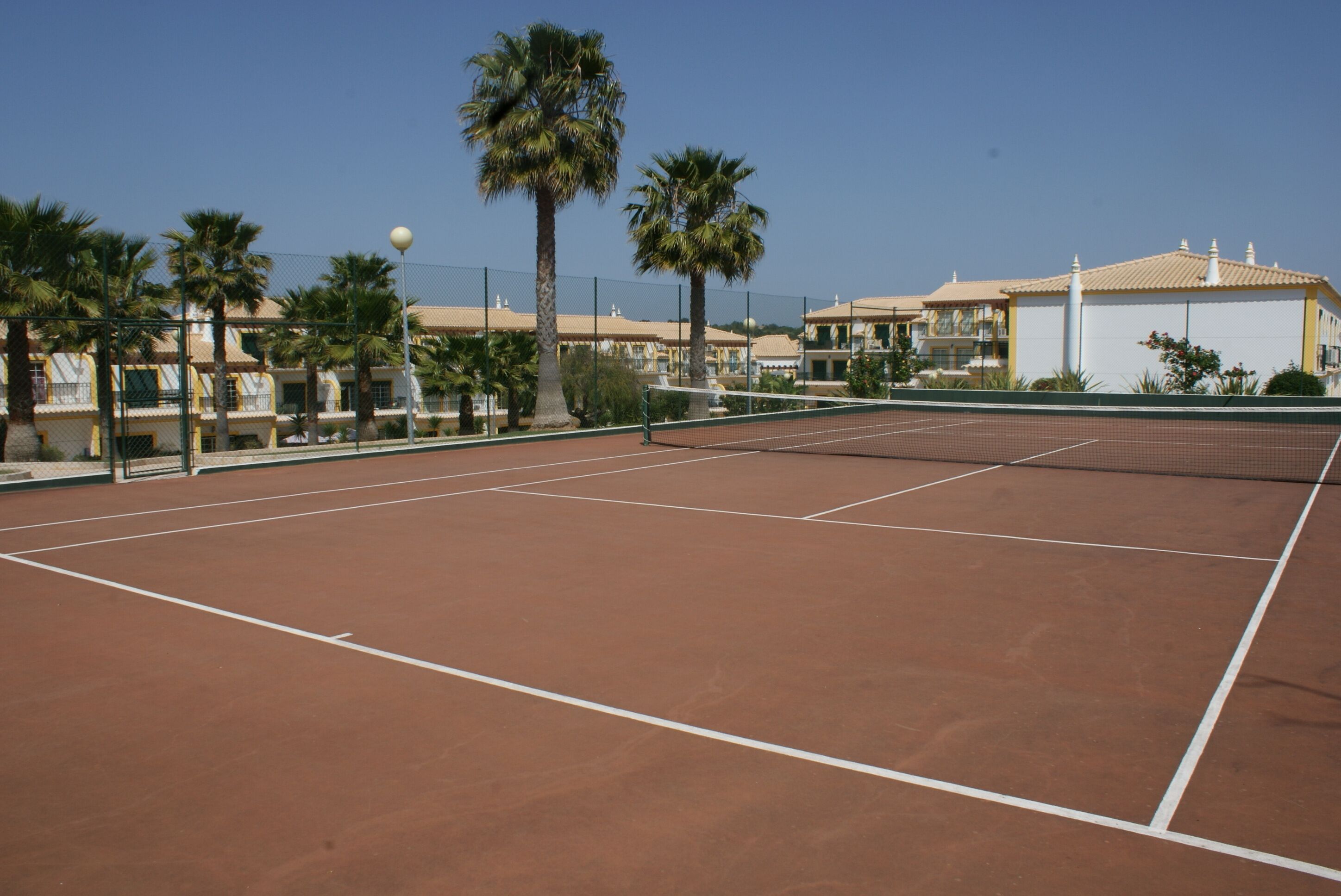 Una cancha de tenis con una red, rodeada de palmeras y un edificio con techo de tejas.