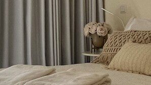 Ein ordentlich gemachtes Bett mit gefalteter Decke, ein Nachttisch mit Blumenvase und graue Vorhänge.