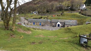A rural setting with a stone building featuring a blue door, surrounded by grassy areas and trees. - Cottage in the Highest Village in Scotland (Wanlockhead)