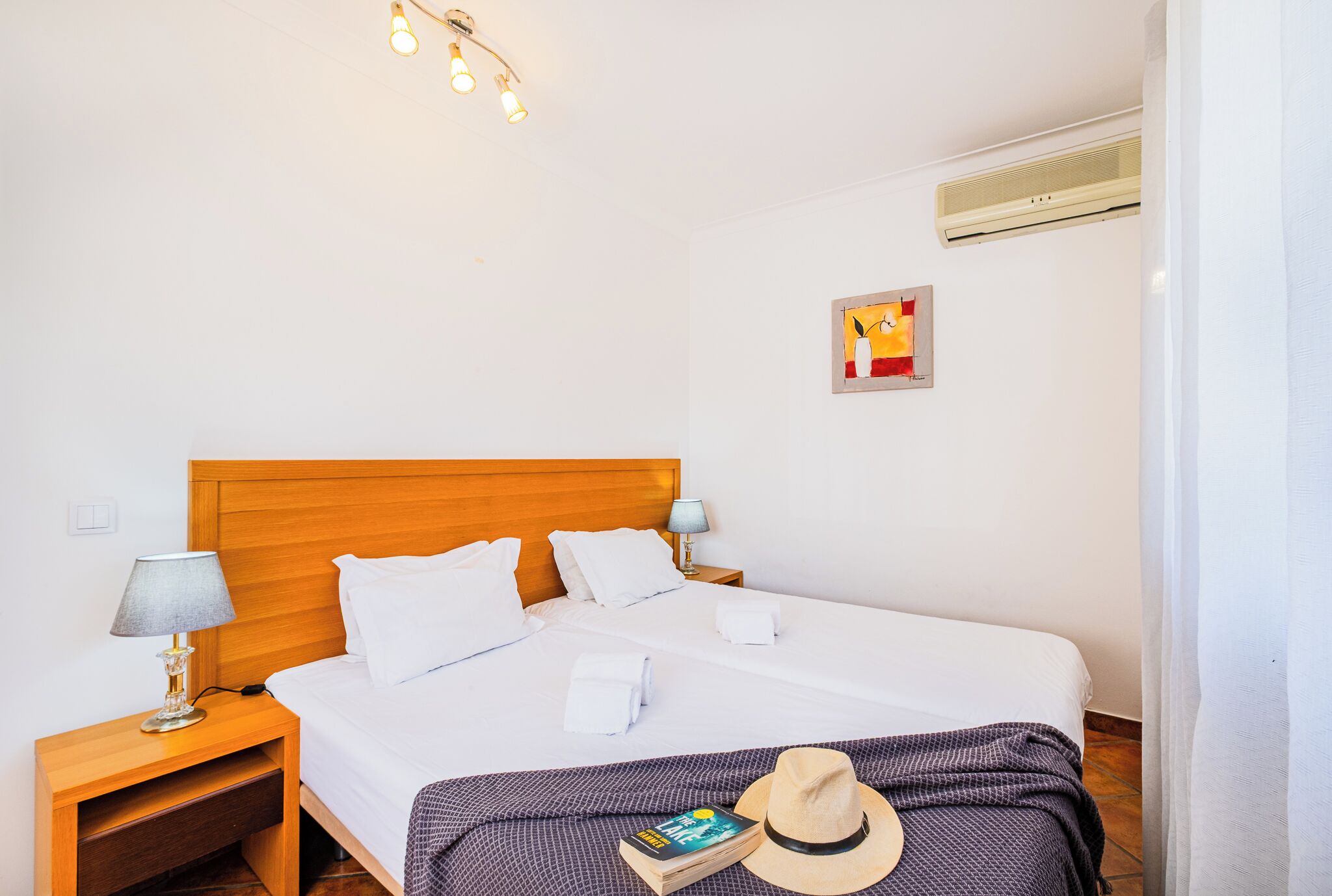 Een net opgemaakt bed met witte lakens, een houten hoofdbord en een bedlampje. Op een klein tafeltje staan een lamp en een boek. Op het bed liggen een hoed en een tijdschrift.