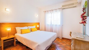A hotel room with a wooden headboard, a bed with white and yellow bedding, a nightstand with a lamp, a window with curtains, and an air conditioning unit. - Cosy Apartment in Jardins Vale de Parra (Albufeira)