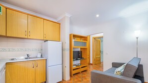 A compact kitchen with wooden cabinets, a white refrigerator, and a tiled backsplash. There is a grey sofa with a patterned cushion in the living area. - Cosy Apartment in Jardins Vale de Parra (Albufeira)
