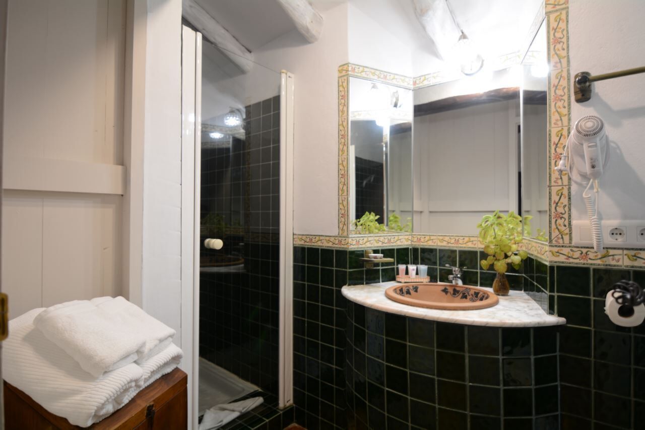 Un baño con pared de azulejos negros, un lavabo blanco con una pila decorativa, un espejo con marco dorado y un perchero con toallas dobladas.