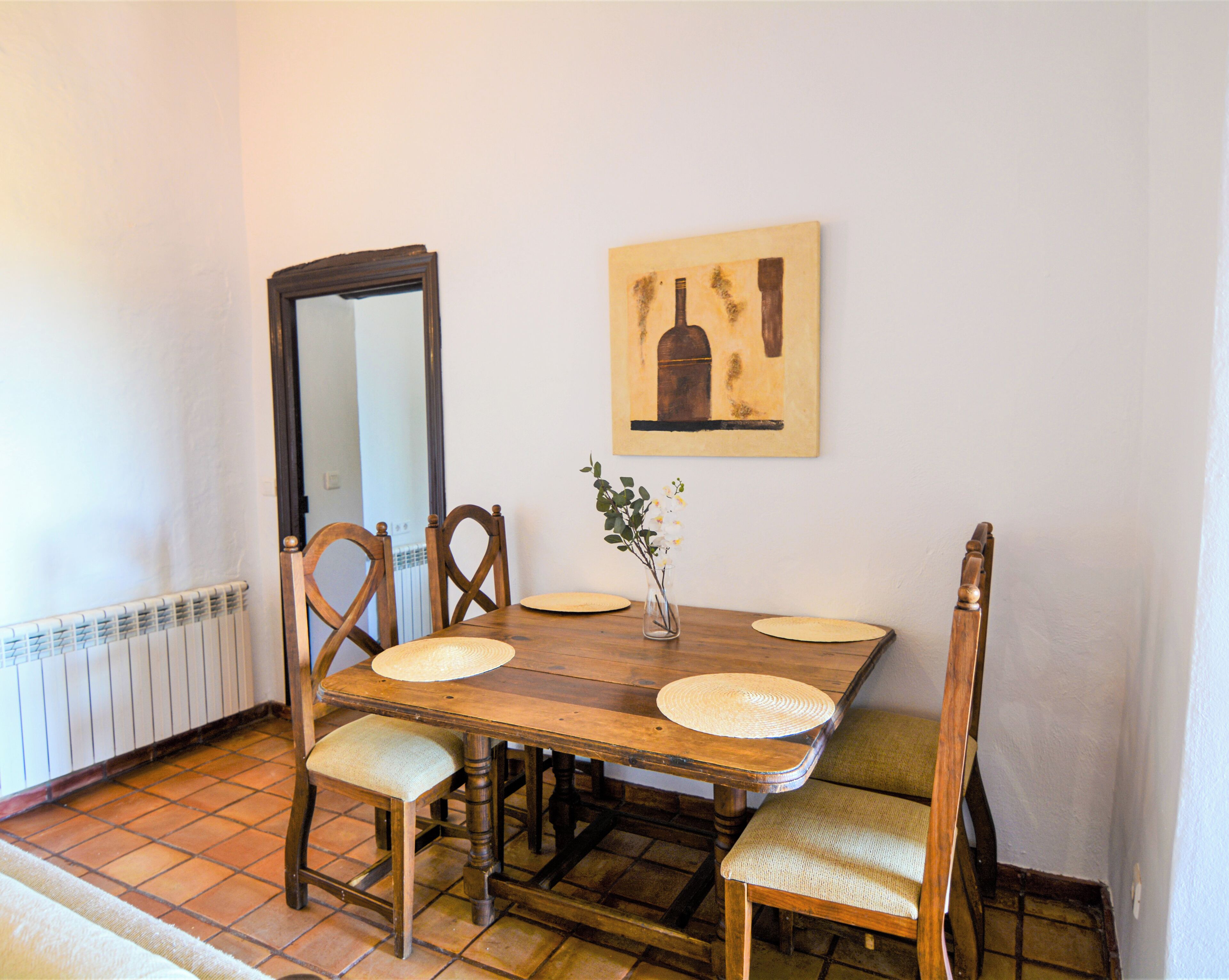 A dining area with a wooden table, four chairs, a vase with flowers, and a painting on the wall.