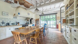 Cucina spaziosa con un grande tavolo e sedie di legno, zoccolo paraschizzi piastrellato e finestra vista su vegetazione.