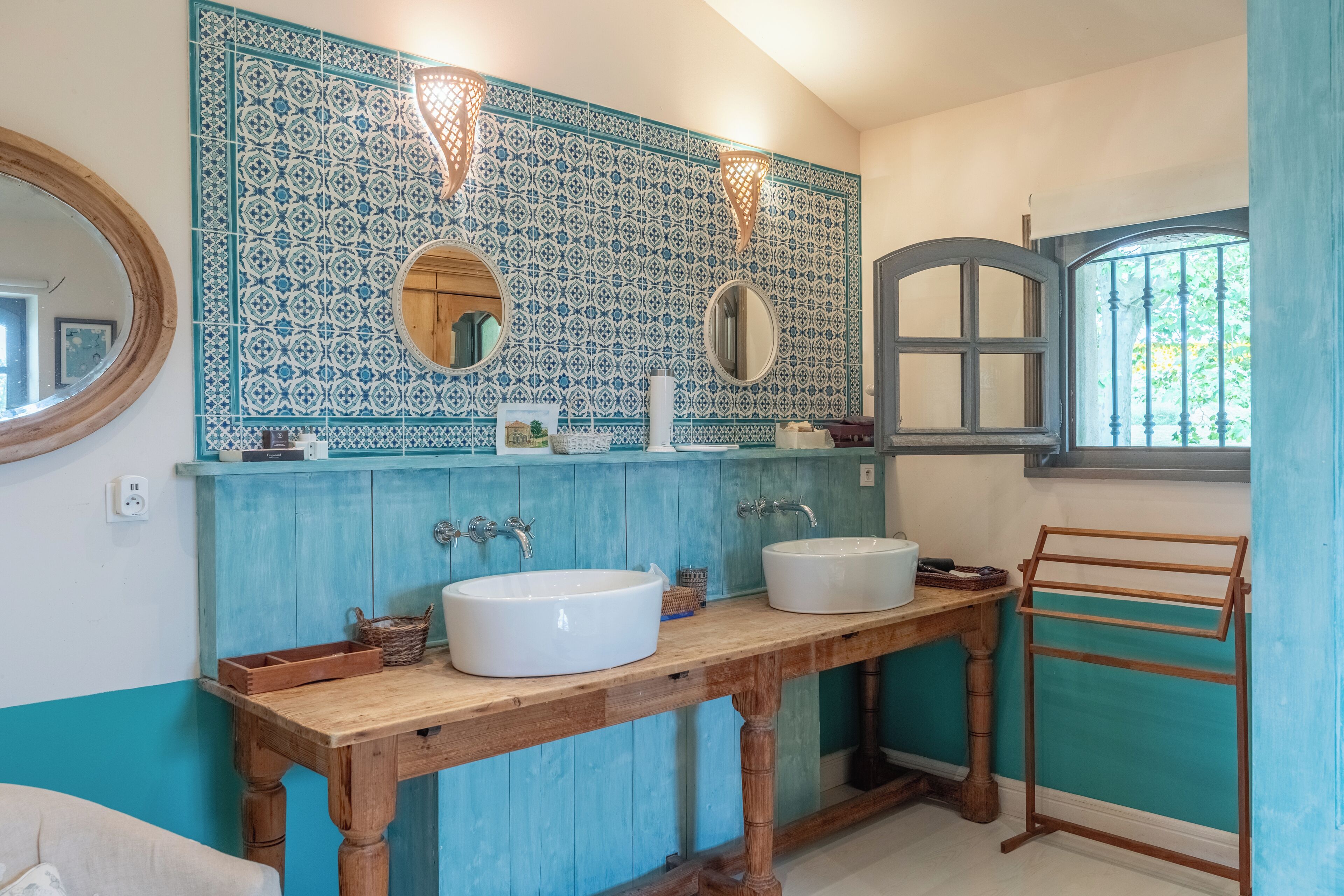 A bathroom with two white sinks, a wooden vanity, a blue and white patterned backsplash, and a window with a view of greenery.
