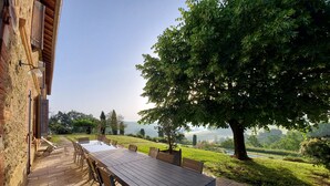 Un comedor con una mesa larga y sillas, suelo de piedra y vistas a un paisaje verde con árboles y colinas.