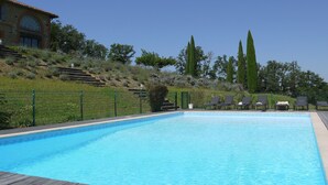 Una piscina rectangular con una cubierta de madera en primer plano, rodeada por una cerca verde y una colina con árboles y un edificio al fondo.