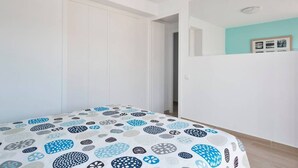 Quarto com cama coberta por um cobre-leito estampado, paredes brancas, uma porta e uma parede azul clara com quadros.