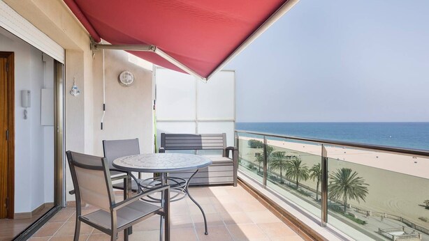 Uma varanda com mesa e cadeiras, um toldo vermelho e vista para a praia e o oceano.