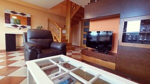 A modern living room with a leather sofa, a glass coffee table, a flat-screen TV, and a staircase.