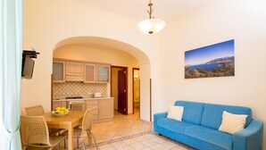 A living room with a blue sofa, a dining table with chairs, a kitchen with cabinets and a tiled backsplash, and a painting of a coastal landscape.
