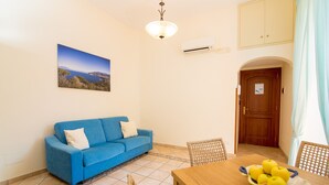 A room with a blue sofa, a dining table with a bowl of apples, a chandelier, and a painting on the wall.