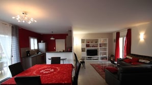 Ein modernes Wohnzimmer mit einem roten Sofa, einem Esstisch mit Stühlen, einer Küchenzeile und einem Fernseher im Hintergrund.