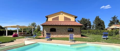 Zona de piscina con edificio de piedra, sillas de descanso y una pérgola.
