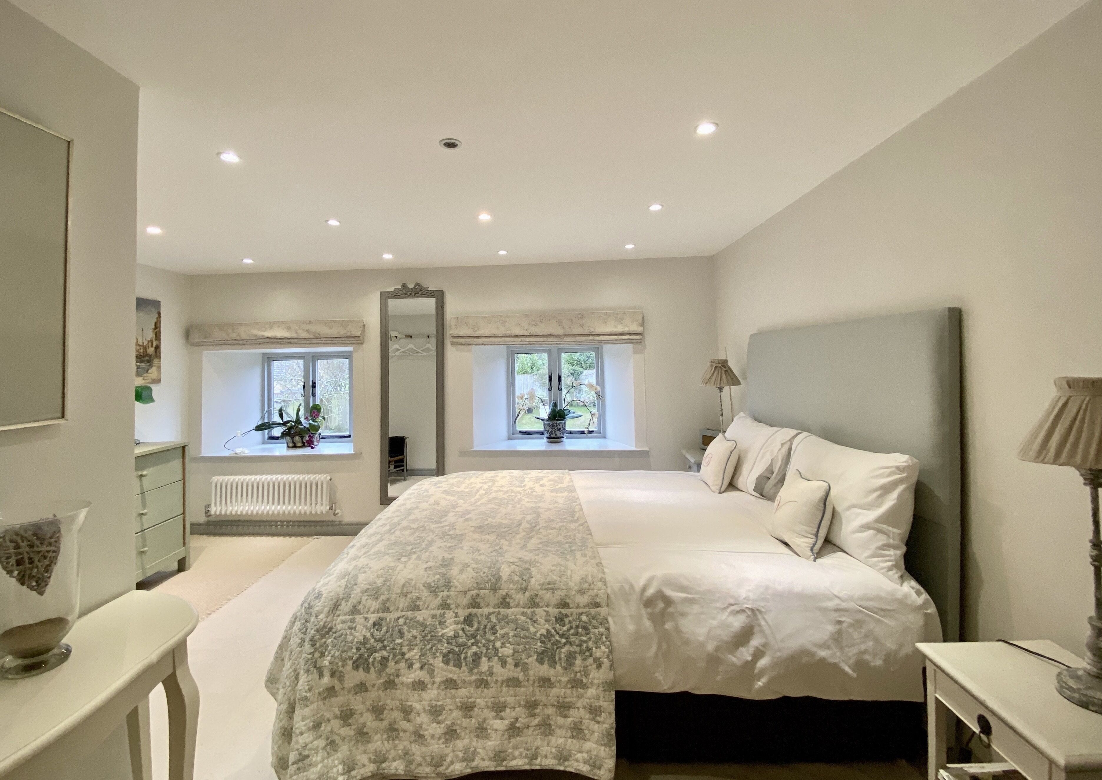 A bedroom with a large bed, two windows with plants, a radiator, and a mirror.