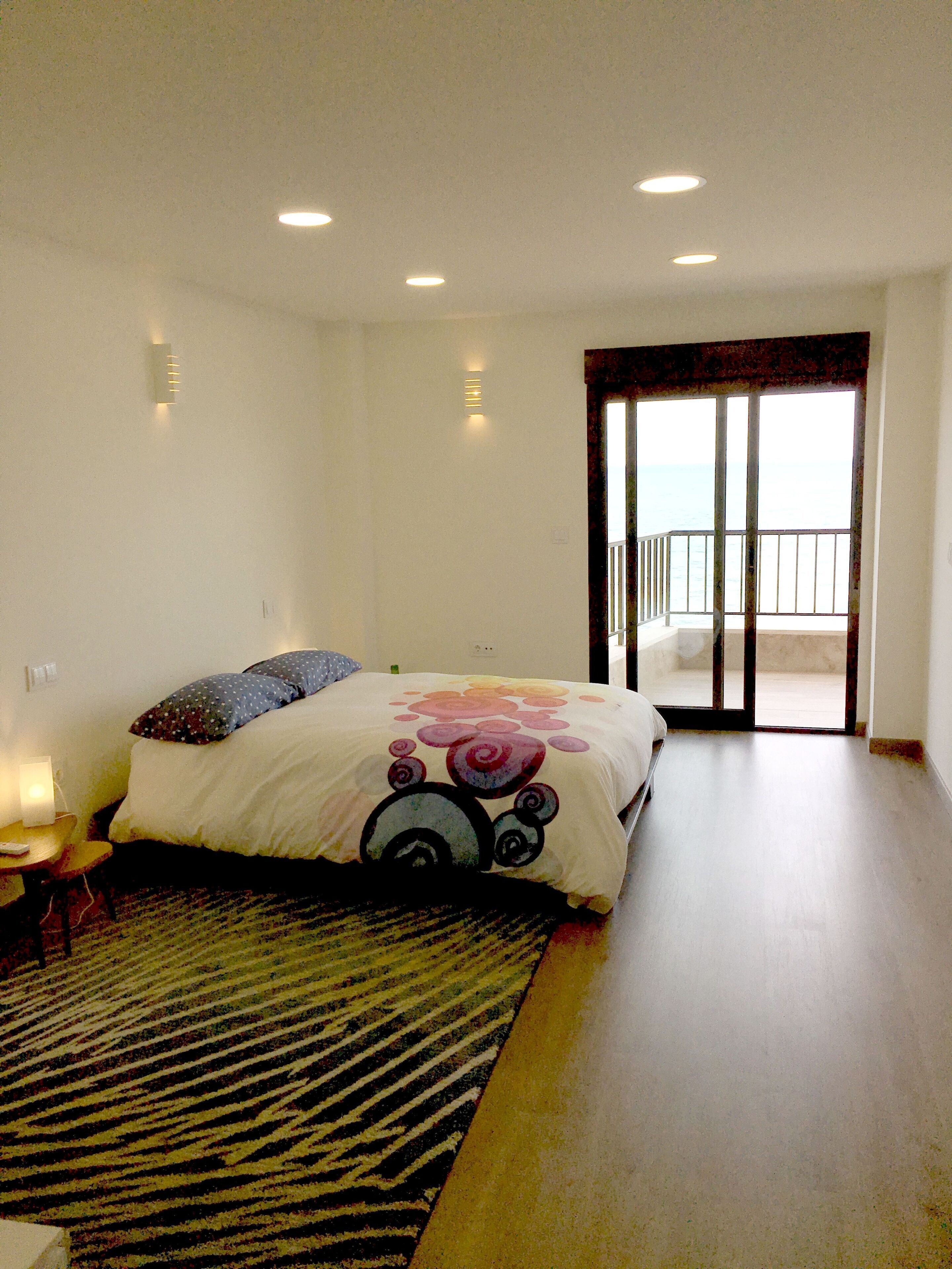 A bedroom with a bed, a nightstand, a lamp, a window with a view, and a door leading to a balcony.