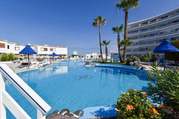 A swimming pool with blue umbrellas, palm trees, and a hotel building in the background.