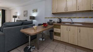 Een knus leefruimte met een kitchenette, een grijze bank, een kleine eettafel met een kopje en fruitschaal, en houten kastjes.