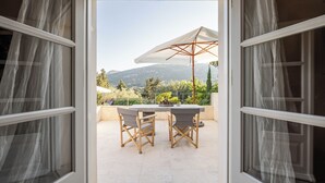 An open door leading to a balcony with a table and chairs, a white umbrella, and a view of mountains and greenery. - Kalliroi - Exquisite Villa Designed Exclusively For Two with Large Private Pool. (Pylaros)